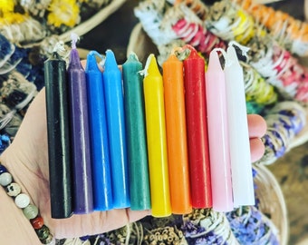 May include: A set of 10 small candles in various colors, including black, purple, blue, green, yellow, orange, red, pink, and white. The candles are arranged in a rainbow pattern and are sitting on a hand.