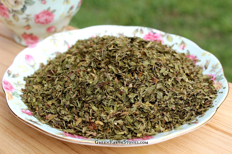 May include: A close-up of a pile of dried green mint leaves on a white and floral patterned plate. The mint leaves are loose and ready to be used for tea or cooking.