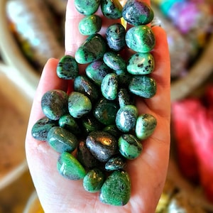 May include: A handful of polished green and black stones with hints of red. A matching beaded bracelet is visible at the bottom. The stones are smooth and rounded, with a glossy finish. The background is blurred, suggesting a natural setting.