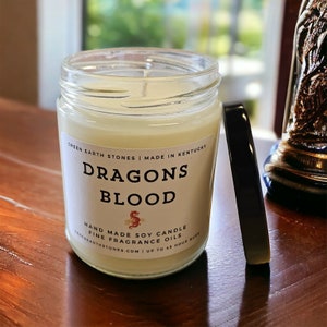 Pu&ograve; includere: Una candela in vetro in un barattolo con un'etichetta bianca che recita "Green Earth Stones | Made in Kentucky" e "Dragons Blood". L'etichetta include anche il testo "Hand Made Soy Candle Fine Fragrance Oils GreenEarthStones.com | Up to 48 Hour Burn".