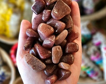May include: A collection of tumbled red and brown stones, also known as red jasper, in a hand. The stones are smooth and polished.