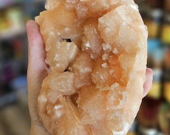 Può includere: Un grande ammasso di cristalli di selenite di colore arancione con una superficie ruvida e testurizzata. Il cristallo è composto da molti cristalli individuali più piccoli che sono raggruppati.