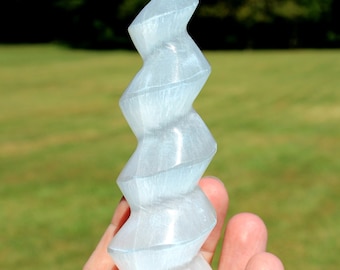 May include: A clear, spiraling selenite crystal sculpture. The polished stone has a zig-zag design and is held in a hand against a blurred green background. The sculpture is approximately 8 inches tall.
