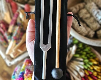 Peut inclure: Un diapason en métal argenté avec le texte 432Hz gravé sur la base, accompagné d'un maillet en bois avec une tête en caoutchouc noir. Les deux objets sont placés sur une pochette en tissu noir à cordon, offrant une vue claire des éléments.
