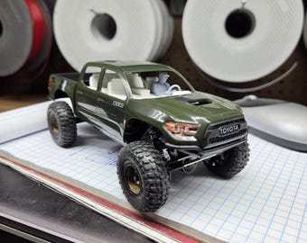May include: An olive green Toyota pickup truck model with large black off-road tires, a white interior, and a small driver figure. The front grille features the Toyota logo, and the side panel displays the SCX10 text in a small white box.