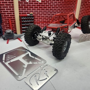 May include: A red and silver model off-road vehicle with large black tires. The vehicle has a roll cage and is displayed with metal plates. The background features a brick wall and a garage setting.
