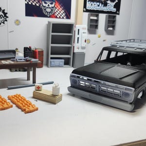 May include: A detailed diorama of a garage scene featuring a black SUV model with a roof rack. The scene includes a workbench with a steering wheel and dashboard, orange traction mats, and other miniature accessories. The walls are decorated with signs.