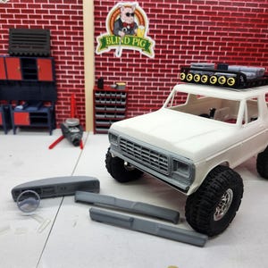 May include: A white toy SUV with black tires and a roof rack is the focal point. Gray accessories are placed in front of the vehicle. The background features a brick wall, cabinets, and a sign that reads "Blind Pig."