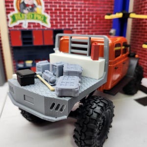 May include: An orange and gray toy truck with large black tires. The truck bed holds several gray miniature objects, including boxes and a hammer. The background features a brick wall and a sign that reads "Blind Pig."