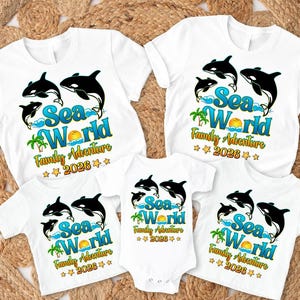 May include: White t-shirts with a colorful "Sea World Family Adventure 2026" design featuring orcas, waves, a sun, and palm trees. The shirts are available in various sizes, including a baby onesie.