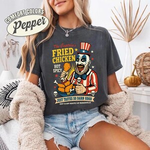 May include: Dark grey t-shirt with a graphic of a clown holding fried chicken. The text on the shirt reads "The Captain's Fried Chicken, Hot Spicy, Just Tastes So Darn Good". The shirt also has the text "Exit 13 off Route 1 at Ruggsville".