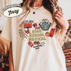 May include: Ivory-colored t-shirt with a heart-shaped graphic featuring basketballs, a basketball hoop, a red dragon, a globe, and the text "EHS High School Musical." The shirt also includes the phrases "Get'cha head in the game" and "breaking free."