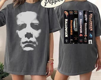 May include: A pepper-colored t-shirt featuring a large white graphic of the Michael Myers mask on the front. The back displays a vertical row of five Halloween VHS tape covers, including Halloween, Halloween II, Halloween III, Halloween 4, and Halloween 5.
