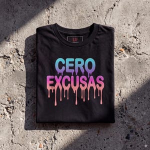 Puede incluir: Camiseta negra con la frase "CERO EXCUSAS" en degradado azul, morado y rosa con efecto goteo. La camiseta está doblada sobre una superficie gris texturizada.
