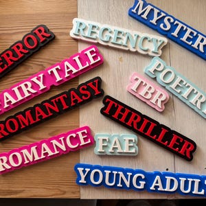May include: A collection of rectangular signs in various colours, each displaying a genre label. The signs read "TERROR," "FAIRY TALE," "ROMANTASY," "ROMANCE," "REGENCY," "MYSTERY," "POETRY," "TBR," "THRILLER," "FAE," and "YOUNG ADULT."