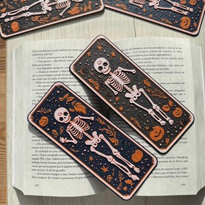 May include: Three Halloween-themed bookmarks featuring a skeleton with a pink skull and orange pumpkins on a black background with white stars. The bookmarks are made of a thick, textured material.