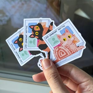 Puede incluir: Tres pegatinas de gato con el texto "VERY IMPORTANT MEETING". Cada pegatina presenta un diseño de gato diferente: un gato negro y naranja, un gato rosa y un gato negro. Las pegatinas se sostienen en una mano.