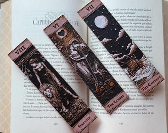Puede incluir: Tres marcapáginas de cartas del tarot con ilustraciones detalladas. Los marcapáginas presentan imágenes de La Fuerza, Los Amantes y El Carro. Cada marcapáginas tiene un borde rosa y números romanos en la parte superior. Los marcapáginas están sobre un libro abierto.