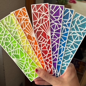 Può includere: Una mano tiene in mano un ventaglio di segnalibri colorati. Ogni segnalibro presenta un design geometrico con un contorno bianco e un diverso riempimento di colore: verde, giallo, arancione, rosso, viola e blu. I segnalibri sono rettangolari con un'estremità a punta.