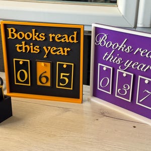 Peut inclure: Deux compteurs de livres décoratifs. L'un est noir avec un cadre orange et le texte "Books read this year" et les chiffres 065. L'autre est violet avec un cadre blanc et le texte "Books read this year" et les chiffres 037.