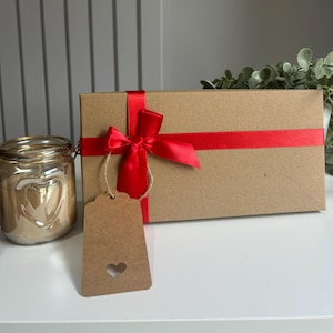May include: A brown rectangular gift box tied with a red satin ribbon and bow. A brown tag with a heart-shaped cutout hangs from the box. A gold jar with a heart design is to the left.