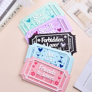 May include: A stack of colorful ticket-shaped cards with various phrases and designs. The cards are in shades of pink, blue, purple, and green, with white text and decorative elements. The text includes phrases like "Friends to Lovers Book Club" and "Forbidden Lover Club."