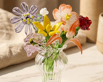 Könnte beinhalten: Eine kleine, klare, gerippte Glasvase mit einem Strauß aus handgefertigten Glasblumen. Das Arrangement besteht aus einem violetten Gänseblümchen, einer leuchtend gelben Blume, einer pfirsichfarbenen Blüte, einer kleinen dunkelroten Rose und hellrosa Blüten.