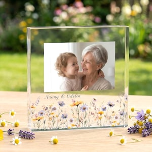 以下が含まれることがあります： 女性と子供の写真が入った透明なアクリルフォトブロック。ブロックは花柄のデザインと「Nanny & Odelia」の文字で装飾されています。 デイジーとラベンダーの花がブロックの周りに配置されています。