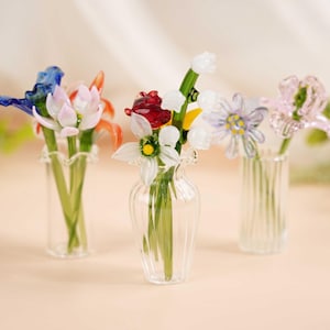 Puede incluir: Tres jarrones de cristal, cada uno con un arreglo diferente de coloridas flores de cristal. Las flores incluyen flores azules, rojas, blancas y rosas con tallos verdes. Los jarrones son transparentes y tienen diferentes formas.