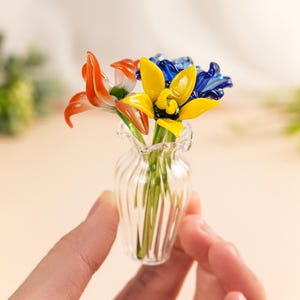 May include: A small glass vase containing colourful glass flowers. The arrangement features orange, yellow, and blue flowers. The vase is clear with a ribbed design. The flowers are held by a person's hand.