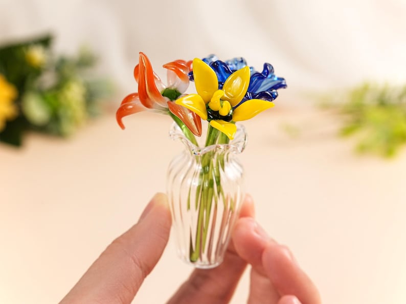 May include: A small glass vase containing colourful glass flowers. The arrangement features orange, yellow, and blue flowers. The vase is clear with a ribbed design. The flowers are held by a person's hand.