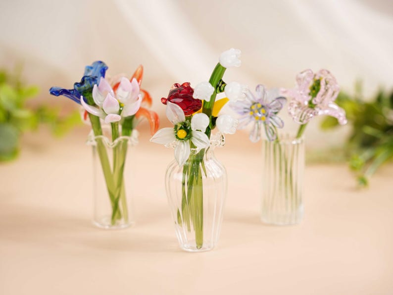 May include: Three glass vases, each containing a different arrangement of colourful glass flowers. The flowers include blue, red, white, and pink blooms with green stems. The vases are clear and have varying shapes, with one having a ruffled edge.