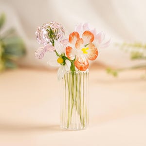 May include: A small glass vase with vertical ridges contains a delicate arrangement of glass flowers. The flowers feature pink, white, and orange petals with yellow centres. The vase is on a light-coloured surface.