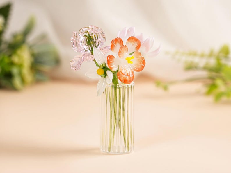 May include: A small glass vase with vertical ridges contains a delicate arrangement of glass flowers. The flowers feature pink, white, and orange petals with yellow centres. The vase is on a light-coloured surface.