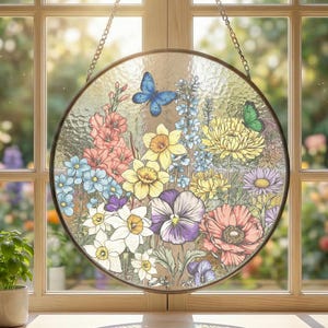 May include: A round stained glass window hanging in front of a window. The glass features a colorful floral design with daffodils, pansies, and butterflies. The window is framed in a dark metal and hangs from a chain.