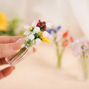 May include: A small glass vase holding a miniature bouquet of flowers. The flowers are white, yellow, red, and blue. The vase has a ribbed design. Other vases with flowers are in the background.