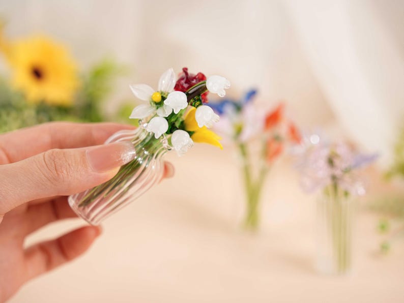 May include: A small glass vase holding a miniature bouquet of flowers. The flowers are white, yellow, red, and blue. The vase has a ribbed design. Other vases with flowers are in the background.