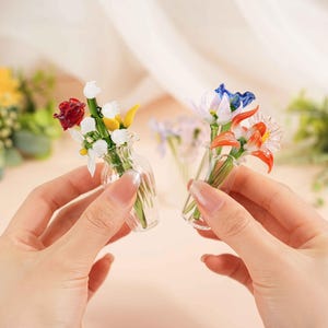 May include: Two miniature glass flower bouquets, each held in a hand. One bouquet features red, white, and yellow flowers, while the other has blue and orange blooms. The clear vases add to the delicate appearance.