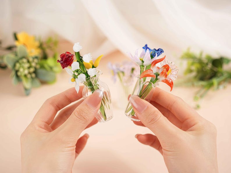 May include: Two miniature glass flower bouquets, each held in a hand. One bouquet features red, white, and yellow flowers, while the other has blue and orange blooms. The clear vases add to the delicate appearance.