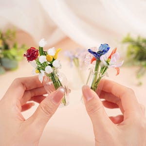 May include: Two miniature glass flower bouquets, each held in a small clear vase. One bouquet features red, white, and yellow flowers, while the other has blue, orange, and pink blooms. The background is a soft, blurred peach colour.