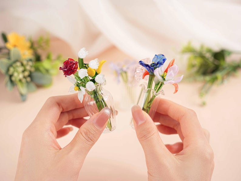 May include: Two miniature glass flower bouquets, each held in a small clear vase. One bouquet features red, white, and yellow flowers, while the other has blue, orange, and pink blooms. The background is a soft, blurred peach colour.