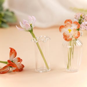 May include: Glass flower sculptures in shades of orange, white, and pink. One flower lies on the surface, while others are displayed in small clear glass vases. The flowers have delicate details and are set against a soft background.
