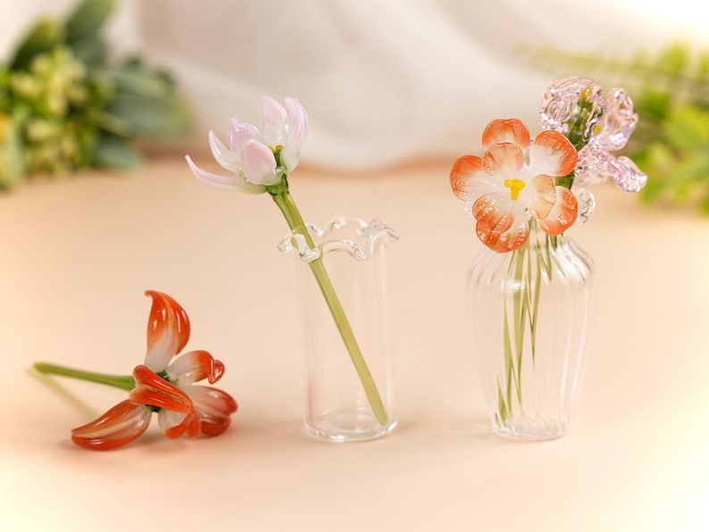 May include: Glass flower sculptures in shades of orange, white, and pink. One flower lies on the surface, while others are displayed in small clear glass vases. The flowers have delicate details and are set against a soft background.