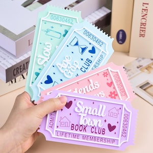 Może przedstawiać: Ręka trzyma cztery kolorowe zakładki do książek w kształcie biletu. Każda zakładka ma unikalny design i tekst, w tym "Slow", "Age Gap Club", "Friends" i "Small Town Book Club". Zakładki są w pastelowych kolorach z elementami dekoracyjnymi.