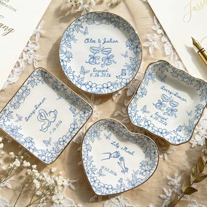 May include: Four white ceramic trinket dishes with gold-toned rims, featuring blue floral and butterfly illustrations. Each dish is personalized with names and dates, including designs of champagne glasses, swans, and hands holding rings.