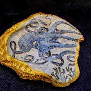 Puede incluir: Concha de ostra decorativa con un diseño de pulpo azul pintado a mano. El pulpo está centrado sobre un fondo crema, con un borde dorado. La concha también presenta coral azul.