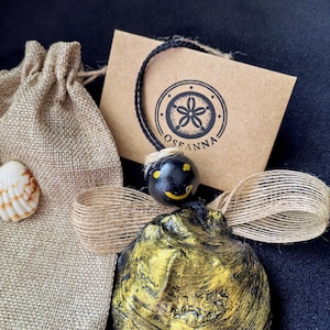 Puede incluir: Una concha decorativa con pintura dorada y negra, una pequeña cuenta negra con una cara sonriente, una bolsa de arpillera y una etiqueta con la palabra "OSFANNA" impresa. Los artículos están dispuestos sobre una superficie oscura.