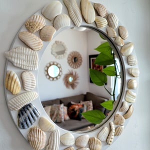 Puede incluir: Un espejo de pared redondo con un marco decorado con un surtido de conchas marinas naturales en varios tonos de blanco, crema y bronceado. Las conchas están dispuestas en un patrón denso y superpuesto alrededor de la superficie de vidrio.