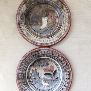 Puede incluir: Dos platos redondos de cerámica con diseños pintados a mano. El plato superior muestra un búho y un cactus. El plato inferior muestra un ave, una planta de hojas y un cactus. Ambos platos tienen bordes color terracota y cenefas geométricas.