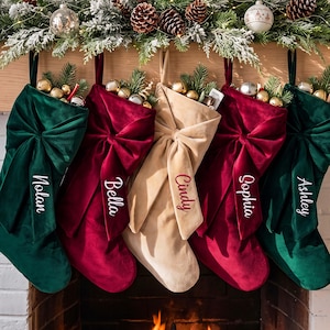 Op de afbeelding: Vijf kerstsokken van fluweel in donkergroen, dieprood en beige, elk voorzien van een grote decoratieve strik en een in witte of rode schrijfletters geborduurde naam. De sokken zijn gevuld met groen en kleine metalen kerstversieringen.